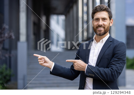 A professional man in business attire presenting or pointing outdoors near modern architecture. Ideal for business, presentation, or success-related projects. 129294348