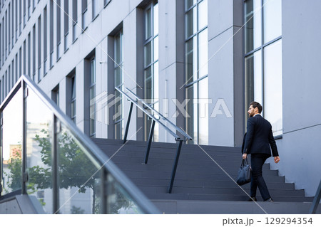 A professional man carrying a bag ascends a flight of stairs beside a contemporary corporate building. The scene symbolizes determination, ambition, and the professional environment 129294354