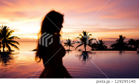 Silhouette of a beautiful woman relaxing in the pool in the setting light of the sun. The pool is surrounded by tropical trees. 129295140
