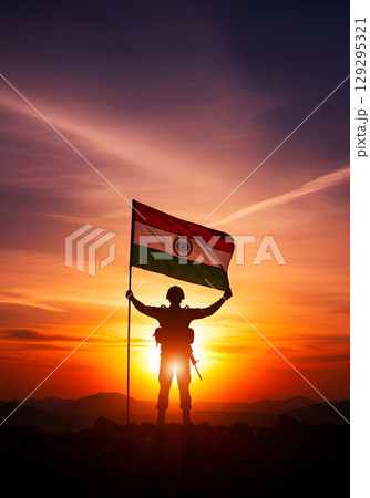 A triumphant Indian soldier, in his 20s, in silhouette, proudly raises the national flag on a mountain peak against a vibrant sunset 129295321
