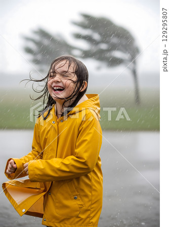 A young female child, joyful and playful, laughs in the rain, wearing a yellow raincoat with a blurred, windswept tree in the background 129295807