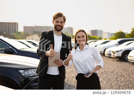 A male car dealer and a woman are standing together, smiling and giving thumbs up in front of a car dealership. 129304678
