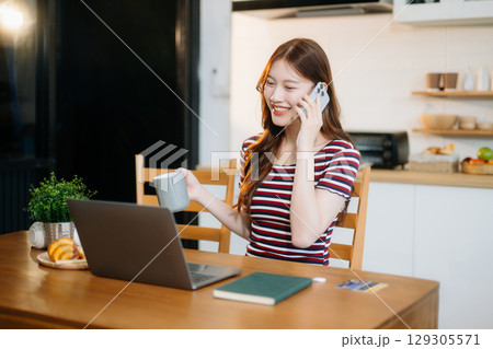 Asian woman working remotely on a laptop and tablet in a cozy kitchen workspace. Perfect for education 129305571