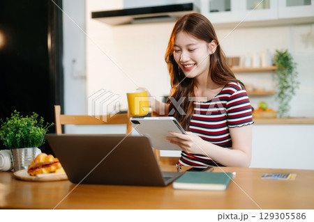 Happy woman with headphones working on a tablet in a bright kitchen. Ideal for themes of remote work Happy woman with headphones working on a tablet in a bright kitchen. Ideal for themes of remote work 129305586