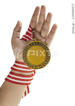Human hand holding golden award medal suspended from a red and white striped ribbon isolated over a white background. Copy space. Symbol of achievement, recognition, or competition 129306417