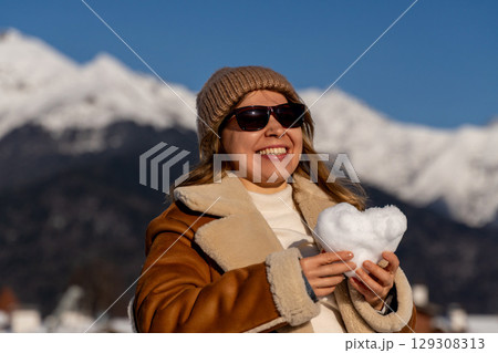 Snow Heart Mountains Woman Winter: Happy tourist holding snow heart, Alps mountains backdrop, sunny winter day. 129308313
