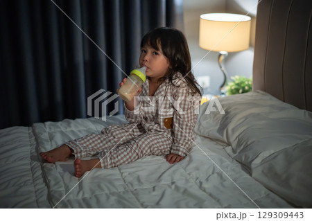 toddler baby girl drinking milk from bottle on a bed at night 129309443