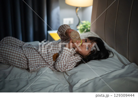 toddler baby girl drinking milk from bottle on a bed at night 129309444