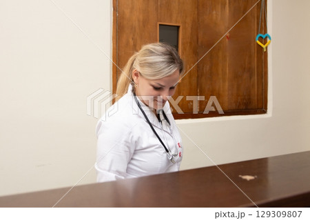 Healthcare professional in a clinical setting wearing a lab coat and stethoscope, likely working at a reception desk or patient check-in. Healthcare professional in a clinical setting wearing a lab coat and stethoscope, likely working at a reception desk or patient check-in. 129309807