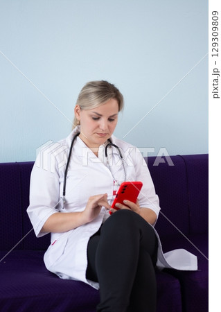 Female doctor in lab coat and stethoscope sitting with legs crossed and using a smartphone, focused and professional. Female doctor in lab coat and stethoscope sitting with legs crossed and using a smartphone, focused and professional. 129309809