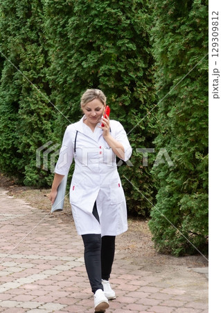 Medical professional in a lab coat walks along a brick path while talking on a cell phone surrounded by greenery on a sunny day. 129309812