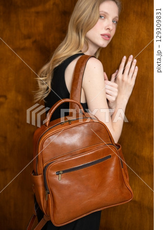A fashion model is captured from a back-facing perspective, holding a rich brown leather backpack and looking away, showcasing the product in a minimalist setting. 129309831