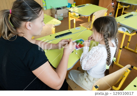 Teacher helping a student with colorful modeling clay at a light green school desk. The student has braids. Classroom environment. Teacher helping a student with colorful modeling clay at a light green school desk. The student has braids. Classroom environment. 129310017