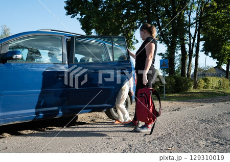 Woman opening a blue car door on a rural road, with another person exiting the vehicle near some trees on a sunny day. Backpack detail. 129310019