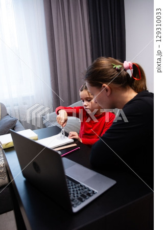 Child learning with tutor at home, using a drawing compass on paper, with a laptop in foreground, cozy, educational setting. Child learning with tutor at home, using a drawing compass on paper, with a laptop in foreground, cozy, educational setting. 129310033