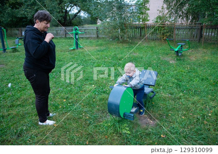 Young child explores outdoor fitness equipment at a park, watched over by an adult, engaging in early physical activity and learning. Young child explores outdoor fitness equipment at a park, watched over by an adult, engaging in early physical activity and learning. 129310099