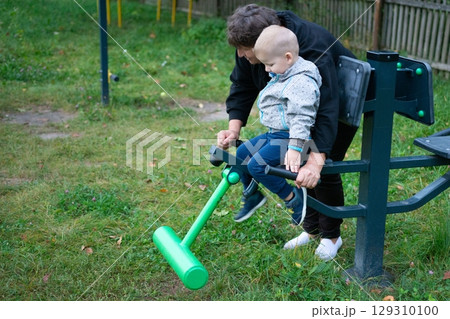 A young child sitting on outdoor fitness equipment with help from an adult in a grassy park on a cloudy day. Learning healthy habits. 129310100