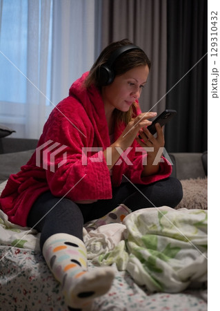 Woman in robe uses a mobile device while sitting on the bed listening to headphones in the bedroom. She seems comfortable and relaxed. 129310432