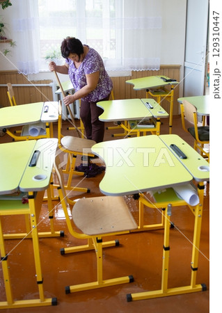 Woman mops a flooded classroom with yellow desks. Water covers floor as she cleans with mop. Light streams through window with lace curtains. Woman mops a flooded classroom with yellow desks. Water covers floor as she cleans with mop. Light streams through window with lace curtains. 129310447