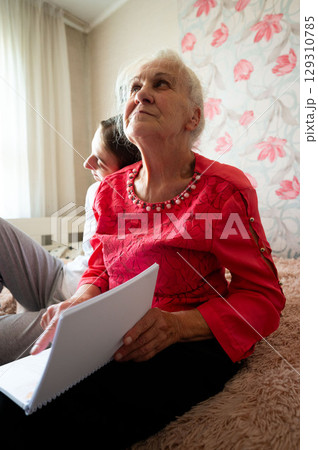 Elderly woman engaging in thoughtful reflection while seated on a cozy couch with a younger woman in a warm, inviting room filled with soft light during a quiet afternoon 129310785