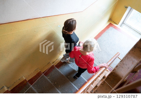 Supportive moments shared on a staircase between a young woman and an elderly lady as they navigate their way together in a bright, inviting space during the day 129310787