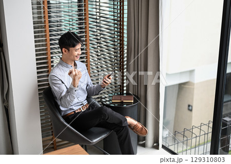 Happy businessman celebrating success while looking at smartphone near office window 129310803