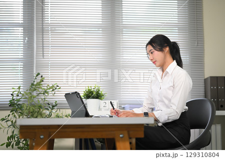 Asian office worker working with digital tablet, taking notes in a bright office 129310804