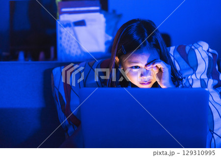 Asian girl sitting on the couch watching laptop at night. Parental control, screen addiction, and bedtime behavior. Asian girl sitting on the couch watching laptop at night. Parental control, screen addiction, and bedtime behavior. 129310995