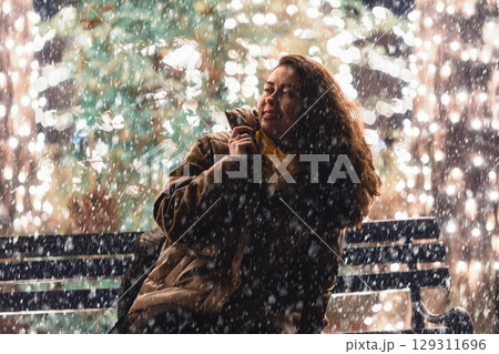 Happy young Caucasian woman in winter clothes sitting on a bench during snowfall. Shiny golden electric garlands in the background. Concept of Christmas and celebration. Happy young Caucasian woman in winter clothes sitting on a bench during snowfall. Shiny golden electric garlands in the background. Concept of Christmas and celebration. 129311696