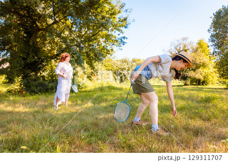 Caucasian mother and daughter dressed in casual clothes laughing while playing badminton in the city park. Family holiday, fitness and connection outdoors. 129311707