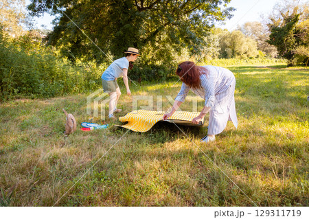 Mature mom and young daughter placing blanket for picnic in green urban park. Parenting time, outdoor activity and weekend relaxation. 129311719