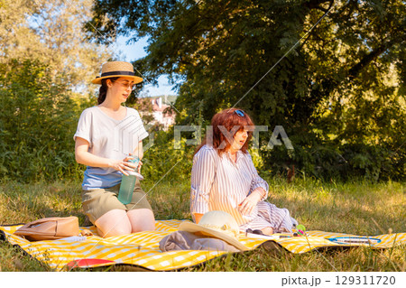 Mature woman and her young daughter spending warm afternoon on a plaid in nature. Concept of family holidays and mental health care. Mature woman and her young daughter spending warm afternoon on a plaid in nature. Concept of family holidays and mental health care. 129311720