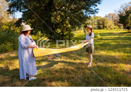 Middle aged mother and teenage Caucasian daughter spread picnic blanket on grass in summer park. Family bonding, vacation time and outdoor leisure concept. 129311725