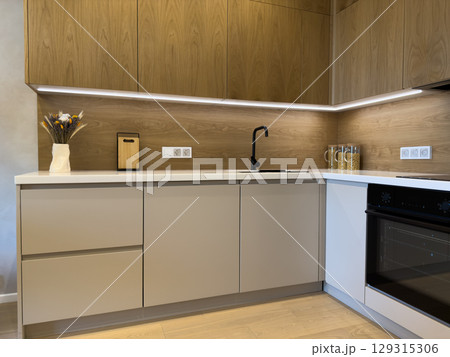 Modern kitchen interior featuring light wood cabinets, sleek white countertops, and contemporary appliances, with a minimalist design highlighted by ambient lighting and small decor items 129315306