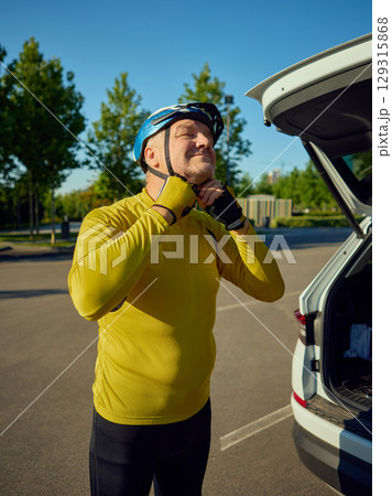 Smiling older cyclist tightening helmet strap before ride beside white car Smiling older cyclist tightening helmet strap before ride beside white car 129315868