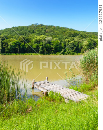 wooden pier for fishing on the lake. summer vacation near the water reserve. landscape with green grass on the shore and forest on the hill. blue sky on a sunny day. calm scenery in the morning wooden pier for fishing on the lake. summer vacation near the water reserve. landscape with green grass on the shore and forest on the hill. blue sky on a sunny day. calm scenery in the morning 129317981