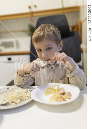 Blond child eating noodles with sausage in the kitchen Blond child eating noodles with sausage in the kitchen 129318001