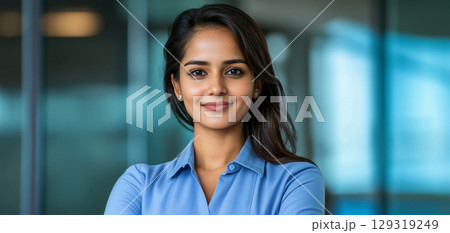Confident woman smiling with arms crossed, standing in modern office environment with large windows and soft natural light during the day 129319249