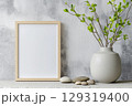 Decorative home setting with an empty frame for poster mockup, neutral tones, and a vase featuring fresh branches on a shelf with smooth stones 129319400