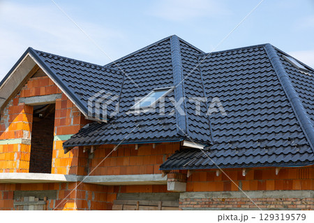 Construction of a house with a black tiled roof and orange brick walls in the framework stage during bright daylight Construction of a house with a black tiled roof and orange brick walls in the framework stage during bright daylight 129319579