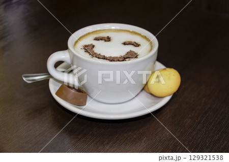 A cup of coffee with candy and biscuits  on dark background.   Smile is depicted on the coffee from the foam. Coffe - smile 129321538