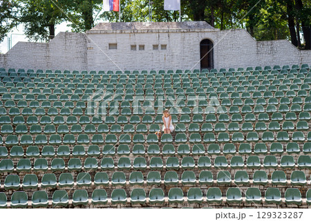Lady wearing a hat is sitting with her dog Cavalier King Charles Spaniel on a seat in a large, open-air stone amphitheater Lady wearing a hat is sitting with her dog Cavalier King Charles Spaniel on a seat in a large, open-air stone amphitheater 129323287