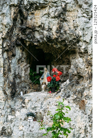 Religious shrine with a Virgin Mary statue and red flowers placed inside a small rock cave. Spiritual devotion, rural Catholicism, sacred natural spaces, cultural heritage Religious shrine with a Virgin Mary statue and red flowers placed inside a small rock cave. Spiritual devotion, rural Catholicism, sacred natural spaces, cultural heritage 129325424