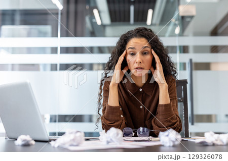 A professional woman sits at her desk surrounded by discarded papers, appearing stressed and frustrated in a modern office setting, suggesting overwhelming workload or creative challenges. A professional woman sits at her desk surrounded by discarded papers, appearing stressed and frustrated in a modern office setting, suggesting overwhelming workload or creative challenges. 129326078