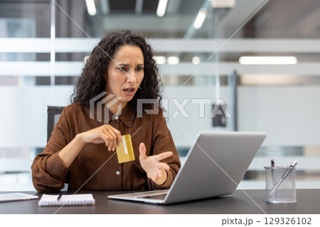 A businesswoman sitting at a desk, expressing frustration holding a credit card and using laptop. The image conveys concepts of online challenges, ecommerce issues, or digital financial difficulties. 129326102