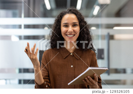 The image shows a professional woman confidently communicating during a business meeting. She holds a notebook, gestures expressively, and smiles warmly, showcasing effective presentation skills 129326105