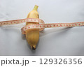A short yellow banana wrapped with a measuring tape, placed on a clean white background. This image highlights both the length and circumference of the fruit. 129326356