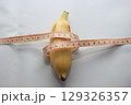 A short yellow banana wrapped with a measuring tape, placed on a clean white background. This image highlights both the length and circumference of the fruit. 129326357