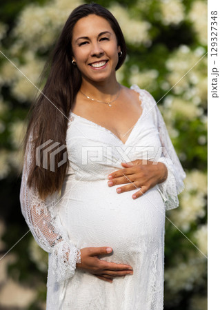 Smiling pregnant woman in lace dress holding her belly in blooming garden Smiling pregnant woman in lace dress holding her belly in blooming garden 129327348