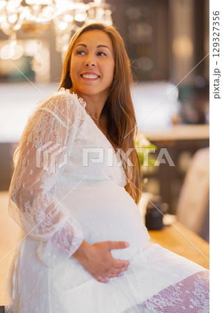 Smiling pregnant woman in elegant white dress indoors, daytime lighting Smiling pregnant woman in elegant white dress indoors, daytime lighting 129327356